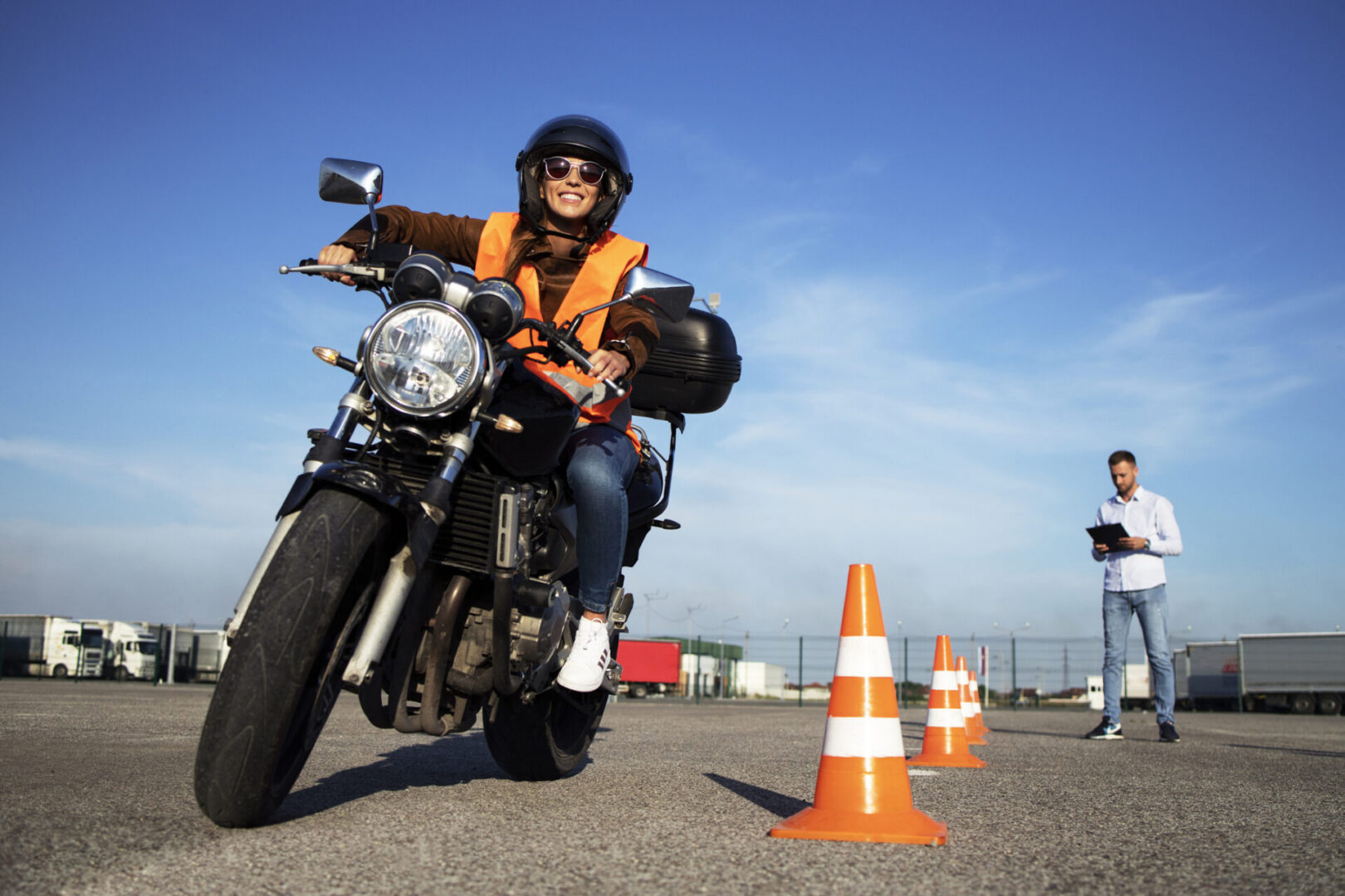Female student with helmet taking motorcycle lessons and practicing ride. In background traffic cones and instructor with checklist rating and evaluating the ride. Motorcycle school of driving.